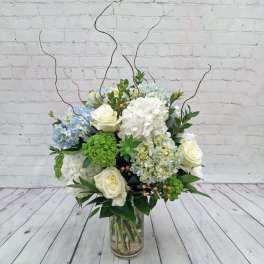Tall glass vase arrangement with white roses, blue and white hydrangeas, green blooms, and curly willow branches.