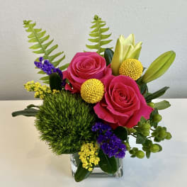 Pink roses in a clear square vase with yellow and purple accents