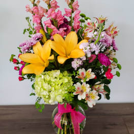Mixed bouquet with yellow lilies, pink snapdragons, and a glass vase tied with a pink ribbon
