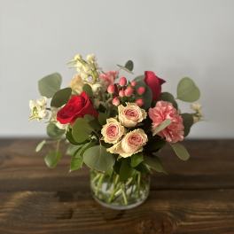 Short arrangement of red roses, blush spray roses, pink carnations, and white flowers in a clear glass vase