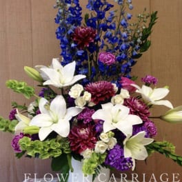 Tall bouquet of white lilies, blue delphinium, and purple blooms in a white vase