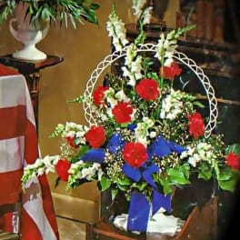 Patriotic red carnation and white snapdragon sympathy arrangement with blue bow in a container.