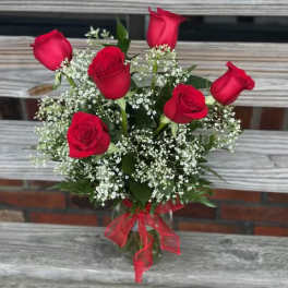 Glass vase of six red roses with white filler flowers and a red bow on a wooden bench