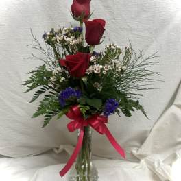 Three red roses in a glass vase with purple filler flowers and a pink ribbon