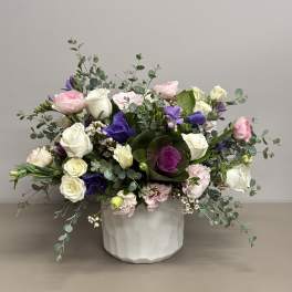 Low arrangement of white roses, pink blooms, and purple flowers in a white ceramic pot