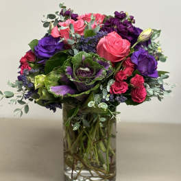 Vibrant arrangement of pink roses, purple flowers, and ornamental cabbage in a clear glass vase