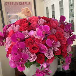 Large arrangement of red and pink roses with magenta orchids in a round white vase.