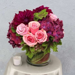 Pink roses and burgundy flowers in a glass vase with a candle beside it