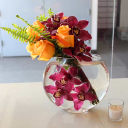 Orange roses and burgundy orchids in a round glass vase