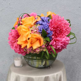Bright bouquet of orange roses, pink dahlias, and purple flowers in a glass bowl vase