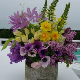 Mixed purple, yellow, and pink flowers arranged in a square stone vase
