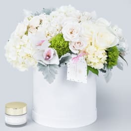 White floral arrangement in a round hat box with a small candle beside it.
