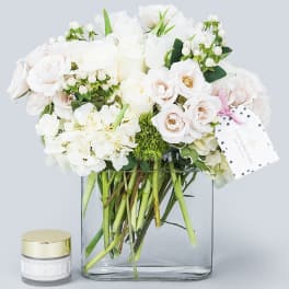 White roses and hydrangeas arranged in a clear glass vase with a small candle jar beside it.