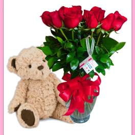 Red roses in a glass vase beside a teddy bear
