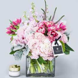 Pink tulips and roses arranged in a square glass vase with a candle beside it.
