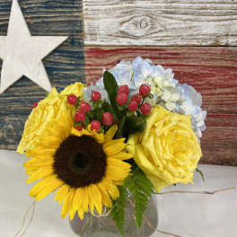 Bouquet of yellow roses, a sunflower, and blue hydrangeas in a glass vase