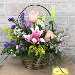 Basket arrangement with pink lilies, roses, and purple flowers beside a yellow candle
