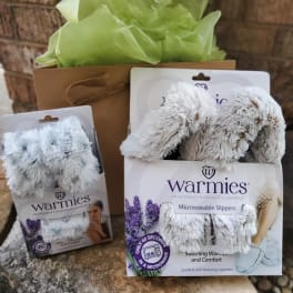 Gift set with two pairs of fuzzy slippers in packaging and a brown bag with green tissue paper