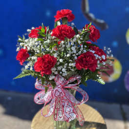 Red carnations in a glass vase with white filler flowers and a heart ribbon