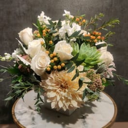White roses and a peach dahlia arranged in a square vase with greenery.