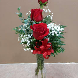 Three red roses in a clear glass vase with baby's breath and a red ribbon