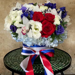 Red, white, and blue floral arrangement in a round box with ribbon