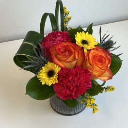 Bright bouquet of orange roses, pink carnations, and yellow daisies in a metal container