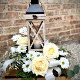 White roses and daisies arranged around a wooden lantern with a candle.