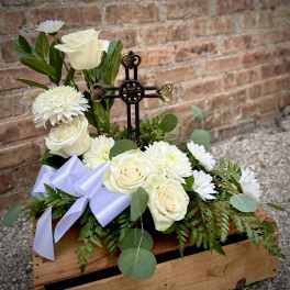 White roses and daisies arranged around a decorative cross in a wooden box