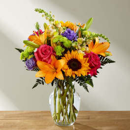 Mixed bouquet of sunflowers, roses, lilies, and purple blooms in a glass vase