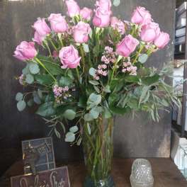 Pink roses arranged in a tall glass vase with eucalyptus and small filler flowers.