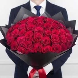 Large bouquet of tightly packed red roses wrapped in black paper, held by a person in a dark suit.