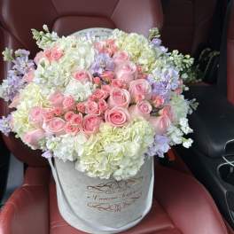 Round hatbox arrangement of pink roses, white hydrangeas, and pastel flowers on a car seat