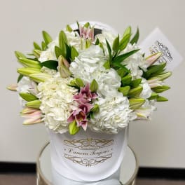 White hydrangeas and pink lilies arranged in a white hatbox with gold script