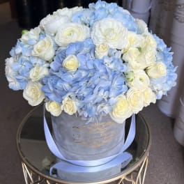 Round hatbox arrangement of white roses and ranunculus with light blue hydrangeas on a mirrored stand