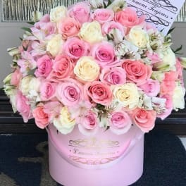 Large round arrangement of pink and cream roses with white accent flowers in a pale pink hat box.
