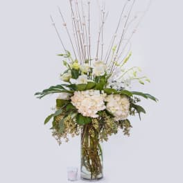 Tall white floral arrangement in a clear glass vase with a small candle beside it.