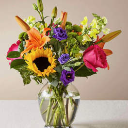 Colorful mixed bouquet in a clear glass vase with a sunflower, roses, lilies, and purple blooms