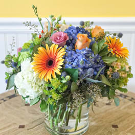 Mixed bouquet in a clear glass vase with blue hydrangeas, roses, and orange gerbera daisies