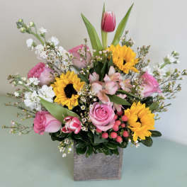 Mixed bouquet with pink roses, sunflowers, and white filler flowers in a square vase