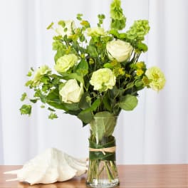 Tall arrangement of pale green roses and carnations in a clear vase wrapped with leaves beside a seashell.