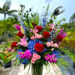 Tall bouquet of red roses, pink lilies, blue hydrangeas, and purple flowers in a glass vase