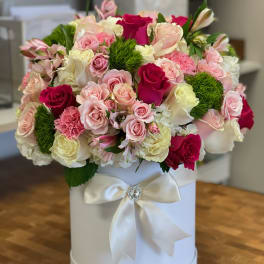 Round arrangement of pink and white roses with green accents in a white hatbox with satin bow