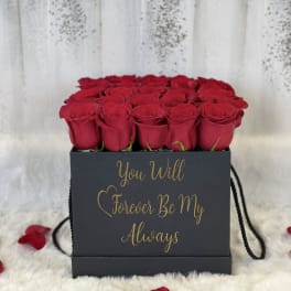 Square black box of red roses with gold script text, surrounded by scattered red rose petals