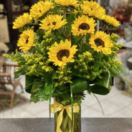 Tall arrangement of yellow sunflowers with yellow filler in a clear glass vase tied with a yellow ribbon.