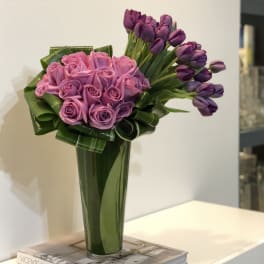 Tall vase arrangement of pink roses and purple tulips with folded green foliage.