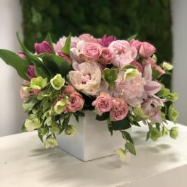 Arrangement of pink roses, peonies, tulips, orchids, and green blooms in a white square vase.