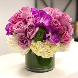 Low round arrangement of mauve roses, purple orchids, and white hydrangeas in a clear glass cylinder vase