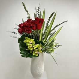 Tall white vase with red roses and green orchids arranged with tall linear foliage.