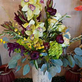 Tall arrangement of green orchids, hydrangea, and dark purple blooms in a white urn-style vase